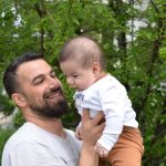 A joyful father lifts his baby in a green, lush outdoor setting.